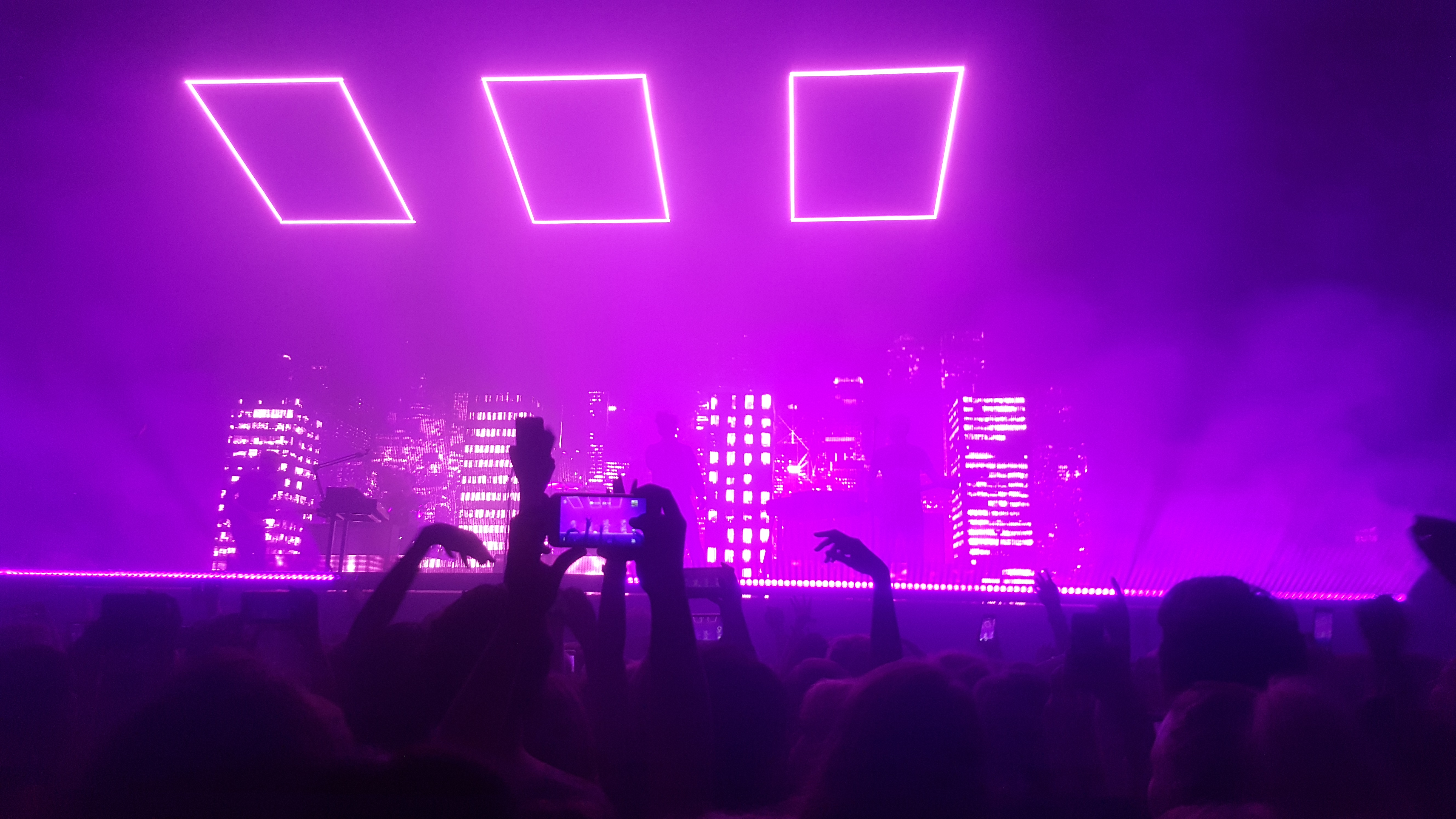 Een afbeelding van een concertzaal met felle paarse lichten en rook.